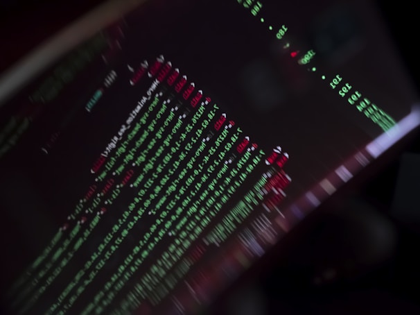 A close-up view of a computer screen displaying programming code with lines of text in green, red, and white against a black background. The code appears to be slightly blurred, creating a moody and abstract visual effect.