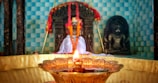 A beautifully decorated temple altar featuring idols draped in colorful garlands. In the foreground, a traditional brass oil lamp emits a warm, glowing light, adding to the sacred ambiance. The background features a tiled wall in shades of blue, adding depth and contrast to the serene atmosphere.