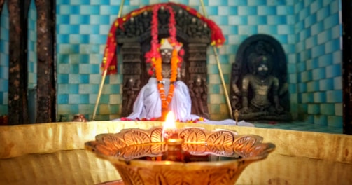 A beautifully decorated temple altar featuring idols draped in colorful garlands. In the foreground, a traditional brass oil lamp emits a warm, glowing light, adding to the sacred ambiance. The background features a tiled wall in shades of blue, adding depth and contrast to the serene atmosphere.