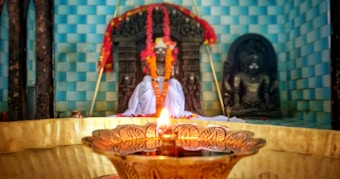 A beautifully decorated temple altar featuring idols draped in colorful garlands. In the foreground, a traditional brass oil lamp emits a warm, glowing light, adding to the sacred ambiance. The background features a tiled wall in shades of blue, adding depth and contrast to the serene atmosphere.