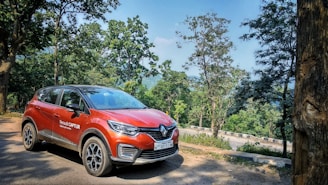 A red Renault Captur car is parked on a dirt road surrounded by lush green trees and hills in the background. The setting suggests a serene, natural environment.