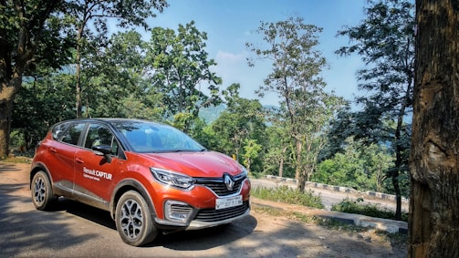 A red Renault Captur car is parked on a dirt road surrounded by lush green trees and hills in the background. The setting suggests a serene, natural environment.
