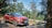 A red Renault Captur car is parked on a dirt road surrounded by lush green trees and hills in the background. The setting suggests a serene, natural environment.