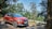 A red Renault Captur car is parked on a dirt road surrounded by lush green trees and hills in the background. The setting suggests a serene, natural environment.