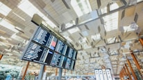 A modern airport terminal interior with high ceilings featuring a geometric pattern and a large digital flight information board displaying departure details. Below, there are illuminated signs and counters with a few travelers milling about. The lighting is bright and airy, contributing to an expansive feel.
