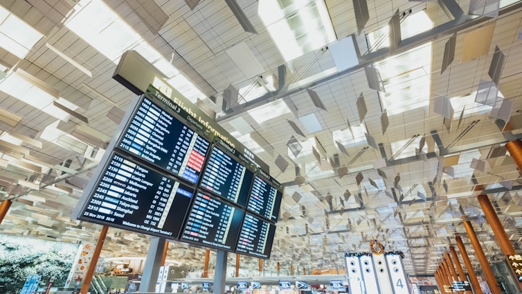 A modern airport terminal interior with high ceilings featuring a geometric pattern and a large digital flight information board displaying departure details. Below, there are illuminated signs and counters with a few travelers milling about. The lighting is bright and airy, contributing to an expansive feel.