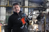 A man is holding a glass sculpture shaped like a horse in a workshop setting. The sculpture features a prominent red color in the body and transparent details on the legs and head. He is wearing glasses and a mustache, dressed in a black jacket, and holding the sculpture with a gloved hand. The workshop has industrial equipment and a rustic interior.