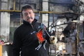 A man is holding a glass sculpture shaped like a horse in a workshop setting. The sculpture features a prominent red color in the body and transparent details on the legs and head. He is wearing glasses and a mustache, dressed in a black jacket, and holding the sculpture with a gloved hand. The workshop has industrial equipment and a rustic interior.