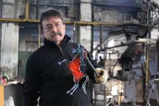 A man is holding a glass sculpture shaped like a horse in a workshop setting. The sculpture features a prominent red color in the body and transparent details on the legs and head. He is wearing glasses and a mustache, dressed in a black jacket, and holding the sculpture with a gloved hand. The workshop has industrial equipment and a rustic interior.