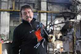 A man is holding a glass sculpture shaped like a horse in a workshop setting. The sculpture features a prominent red color in the body and transparent details on the legs and head. He is wearing glasses and a mustache, dressed in a black jacket, and holding the sculpture with a gloved hand. The workshop has industrial equipment and a rustic interior.