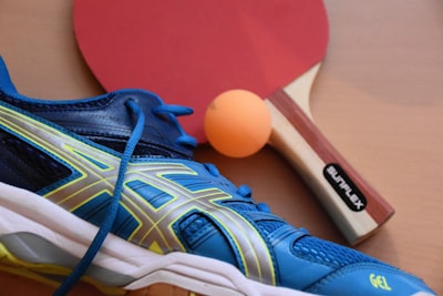 blue and black asics running shoes near ping pong paddle and ball table tennis zoom background