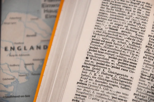 An open dictionary page with text in a foreign language, possibly German, showing various terms and their definitions. Next to it, a blurred map partially visible with the word 'ENGLAND' and parts of the coastline.