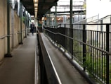A narrow pathway flanked by metal railings on both sides, with a person walking towards the end of the walkway. The pathway is elevated and seems to be leading to a building with multiple lights visible in the background. There is a mix of concrete and greenery along the sides, with a roof structure overhead providing partial cover.
