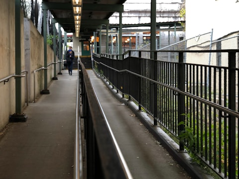A narrow pathway flanked by metal railings on both sides, with a person walking towards the end of the walkway. The pathway is elevated and seems to be leading to a building with multiple lights visible in the background. There is a mix of concrete and greenery along the sides, with a roof structure overhead providing partial cover.