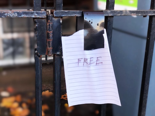 A piece of lined paper with the word 'FREE' written on it is taped to a black metal gate. The tape used is black, and the setting appears to be outdoors with some colorful background blur.