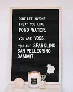 A black felt letter board with a motivational message written in white letters, accompanied by a lit scented candle, a small vase with white flowers, and a wooden object placed on a white surface.