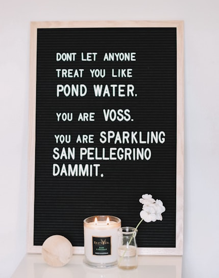 A black felt letter board with a motivational message written in white letters, accompanied by a lit scented candle, a small vase with white flowers, and a wooden object placed on a white surface.
