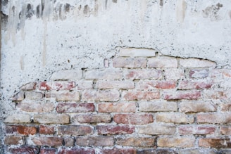 A partially exposed brick wall with an uneven plaster coating. The bricks are of various shades, predominantly red and beige, giving the wall a rustic appearance. The overlying plaster is weathered and has drip marks, adding to the aged look.