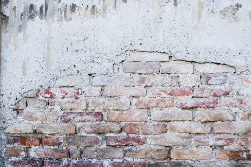 A partially exposed brick wall with an uneven plaster coating. The bricks are of various shades, predominantly red and beige, giving the wall a rustic appearance. The overlying plaster is weathered and has drip marks, adding to the aged look.