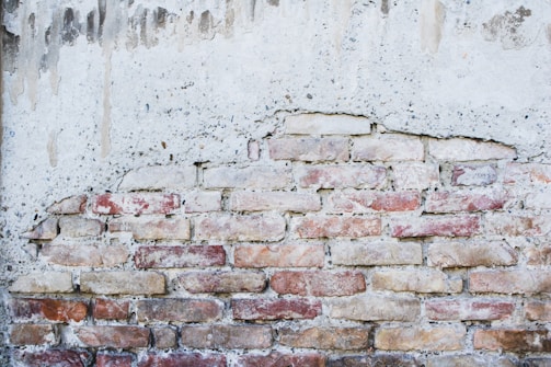 A partially exposed brick wall with an uneven plaster coating. The bricks are of various shades, predominantly red and beige, giving the wall a rustic appearance. The overlying plaster is weathered and has drip marks, adding to the aged look.