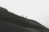 Silhouettes of two hikers and a dog walking up a steep, grassy hill under an overcast sky. The scene conveys a sense of adventure and challenge amidst a minimalist landscape.