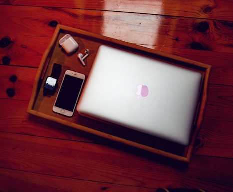 A wooden tray on a wooden floor holds a closed laptop, a smartphone, a smartwatch, a pair of wireless earbuds, and their charging case. The laptop and phone both have a sleek design, suggesting modern technology.