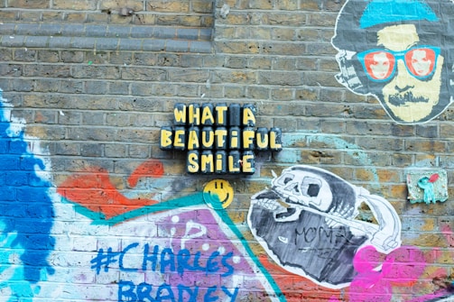 A brick wall is covered with vibrant street art displaying various elements. The phrase 'What a beautiful smile' is prominently featured in bold, yellow letters on a black background. To the right, a stylized face wearing glasses with images inside the lenses is depicted. Below, a drawing of a skeleton holding a scythe labeled 'MOMMA SAID' is visible. Various shapes and splashes of color, including blue, red, and purple, are spray-painted around the wall. The hashtag '#CHARLES BRADLEY' is also part of the mural.
