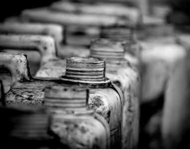 A grayscale close-up view of several weathered and dusty jerry cans lined up in a row, with a focus on their threaded spouts and handles. The image captures the textures and aged surfaces, creating a sense of industrial or rustic setting.