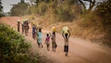 A group of children walking along a dirt road in a rural setting, carrying large yellow and white containers, likely for fetching water. The road is lined with dry grass and sparse trees, suggesting a dry season. The children appear to be smiling and engaged in conversation.