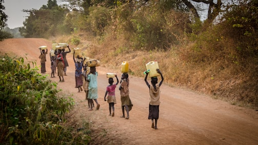 A group of children walking along a dirt road in a rural setting, carrying large yellow and white containers, likely for fetching water. The road is lined with dry grass and sparse trees, suggesting a dry season. The children appear to be smiling and engaged in conversation.