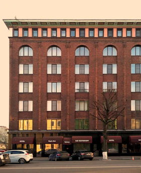 A multi-story brick building with arched windows and several ground floor entrances labeled as a hotel, night club, and bar restaurant. The facade is brown with a contrasting roofline and multiple identical windows. Several cars are parked in front of the building along the street.