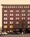 A multi-story brick building with arched windows and several ground floor entrances labeled as a hotel, night club, and bar restaurant. The facade is brown with a contrasting roofline and multiple identical windows. Several cars are parked in front of the building along the street.