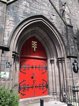A large, arched doorway set within a stone building features striking, red double doors adorned with intricate black wrought iron designs. Above the doors, there is a unique gold symbol. The stone facade is textured and weathered, and signage indicating wheelchair accessibility and other information is mounted on the stone wall. A small patch of greenery can be seen on the lower left side.