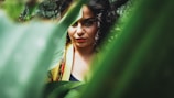 A woman with curly hair is partially visible, framed by lush green leaves that dominate the foreground. Her expression is focused and contemplative, with a hint of mystery due to the foliage obscuring part of her face. She is wearing a colorful garment that adds vibrancy to the natural setting.