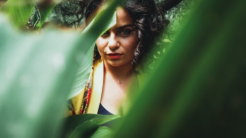 A woman with curly hair is partially visible, framed by lush green leaves that dominate the foreground. Her expression is focused and contemplative, with a hint of mystery due to the foliage obscuring part of her face. She is wearing a colorful garment that adds vibrancy to the natural setting.