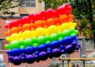 A vibrant arrangement of colorful balloons forming a rainbow pattern, floating outdoors against an urban background with brick walls and trees.