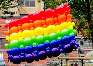 A vibrant arrangement of colorful balloons forming a rainbow pattern, floating outdoors against an urban background with brick walls and trees.