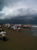 A group of people dressed in ceremonial attire gathers on a beach, preparing or celebrating around boats. The sky is filled with dark, ominous clouds, suggesting an impending storm. The scene is bustling with activity as individuals move around, likely participating in a traditional or cultural event.