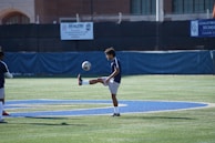 A soccer player is practicing on a field, keeping the ball balanced on his foot. The field is marked with a large painted blue letter, and there are buildings and barriers in the background. The player is wearing a dark blue jersey and white shorts.