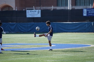 A soccer player is practicing on a field, keeping the ball balanced on his foot. The field is marked with a large painted blue letter, and there are buildings and barriers in the background. The player is wearing a dark blue jersey and white shorts.
