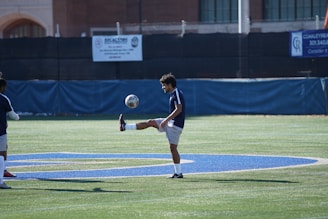 A soccer player is practicing on a field, keeping the ball balanced on his foot. The field is marked with a large painted blue letter, and there are buildings and barriers in the background. The player is wearing a dark blue jersey and white shorts.