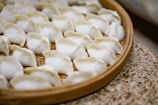 A bamboo steamer tray filled with neatly arranged, uncooked dumplings. The dumplings have a white, slightly translucent appearance, with folds at the top indicating they are freshly handmade.