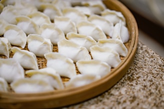 A bamboo steamer tray filled with neatly arranged, uncooked dumplings. The dumplings have a white, slightly translucent appearance, with folds at the top indicating they are freshly handmade.
