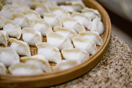 A bamboo steamer tray filled with neatly arranged, uncooked dumplings. The dumplings have a white, slightly translucent appearance, with folds at the top indicating they are freshly handmade.