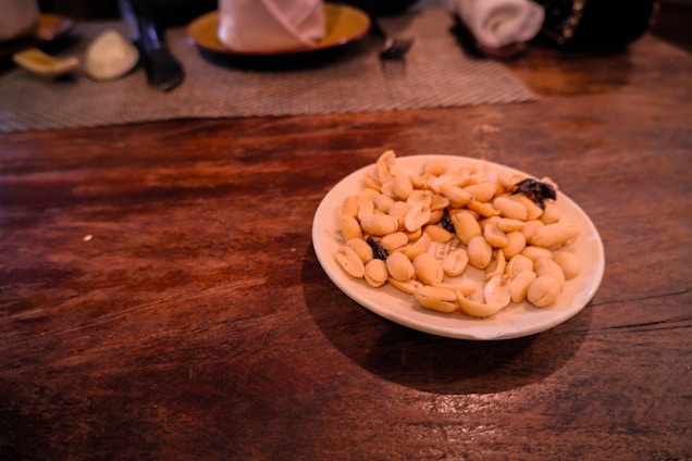 A white plate filled with shelled peanuts and a few raisins is placed on a wooden table, with blurred dining items in the background.