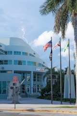 A police department building with a modern design is accompanied by three flags at half-mast in the foreground. A lightning bolt is visible in the sky, adding a dramatic element. Palm trees frame the scene, suggesting a tropical location. A sculpture of an anchor and a sign are also present in the vicinity.