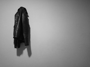 A black leather jacket hangs on a plain light gray wall, casting a shadow. The jacket has stitched patterns and zippers on the sleeves, indicating a stylish and rugged design.