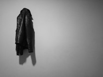 A black leather jacket hangs on a plain light gray wall, casting a shadow. The jacket has stitched patterns and zippers on the sleeves, indicating a stylish and rugged design.
