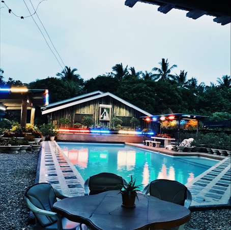 A serene poolside setting with a wooden house in the background, colorful lights decorating the area, and palm trees silhouetted against the evening sky. The pool reflects the vibrant lights, and there are potted plants lining the space. A table with chairs and a small plant is situated in the foreground.