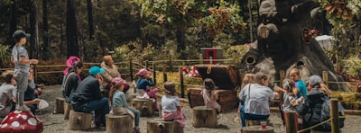A group of children and adults are seated on tree stumps in an outdoor forest setting, listening attentively to a storyteller or performer. They are gathered around an area with a large, whimsical tree sculpture with a face, which appears to be part of a magical or fantasy-themed environment. The scene is surrounded by lush greenery and playful decorations like giant mushrooms.
